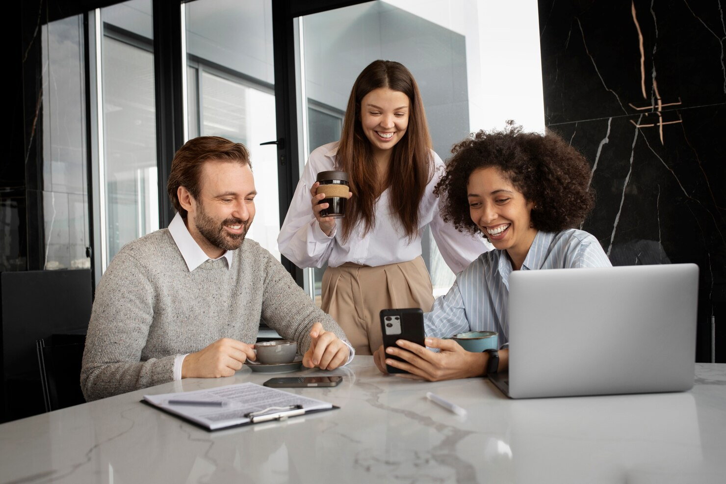 Three employees gather at a table with a laptop and a phone viewing the phone screen and smiling.